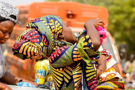OUIDAH, BENIN - Jan 10, 2017: Unidentified Beninese woman in colored clothes carries her baby at the local market. Benin people suffer of poverty due to the bad economyのeditorial素材