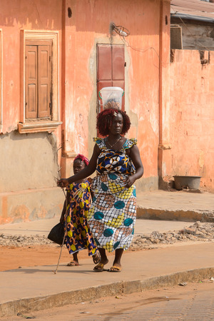 OUIDAH, BENIN - Jan 10, 2017: Unidentified Beninese curly woman in colored clothes on the street with bucket on her head. Benin people suffer of poverty due to the bad economyのeditorial素材