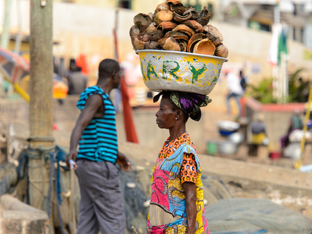 ELMINA, GHANA -JAN 18, 2017: Unidentified  Ghanaian woman in colored dress carries a basin on her...