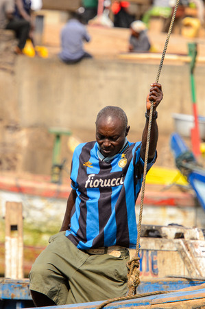 ELMINA, GHANA -JAN 18, 2017: Unidentified  Ghanaian man in striped shirt holds on the rope on the boat in Elmina port. People of Ghana suffer of poverty due to the bad economyのeditorial素材