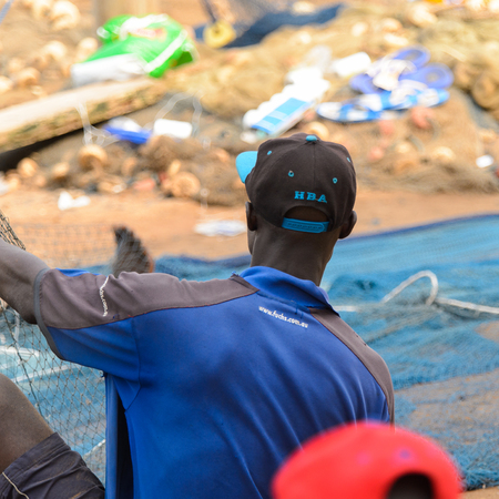 ELMINA, GHANA -JAN 18, 2017: Unidentified  Ghanaian man in blue shirt from behind sits on fishing net in Elmina port. People of Ghana suffer of poverty due to the bad economyのeditorial素材