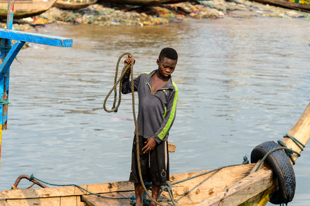 ELMINA, GHANA -JAN 18, 2017: Unidentified  Ghanaian boy holds a rope on the boat in Elmina port. Children of Ghana suffer of poverty due to the bad economyのeditorial素材