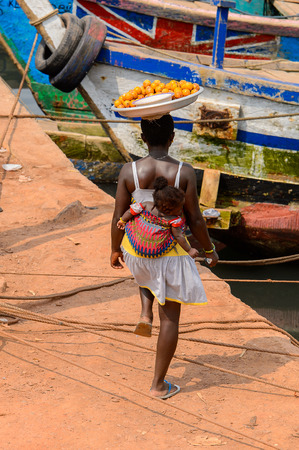 ELMINA, GHANA -JAN 18, 2017: Unidentified  Ghanaian woman carries a baby and a tray on the shore of Elmina port. People of Ghana suffer of poverty due to the bad economyのeditorial素材