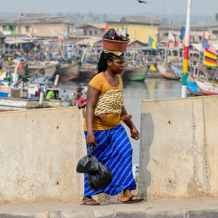 ELMINA, GHANA -JAN 18, 2017: Unidentified  Ghanaian woman carries a baby and a basin on the road of Elmina. People of Ghana suffer of poverty due to the bad economyのeditorial素材