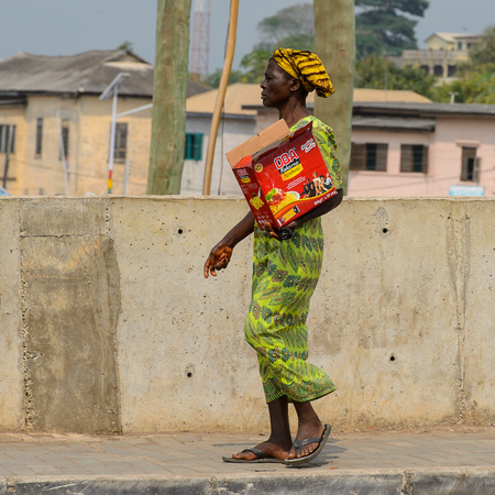 ELMINA, GHANA -JAN 18, 2017: Unidentified  Ghanaian woman walks with a box near the fence in Elmina. People of Ghana suffer of poverty due to the bad economyのeditorial素材