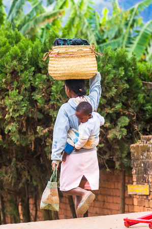 ANTANANARIVO, MADAGASCAR - JUNE 30, 2011: Unidentified Madagascar woman carries her little baby on her back and a bag on her head. People in Madagascar suffer of poverty due to the slow development of the countryのeditorial素材