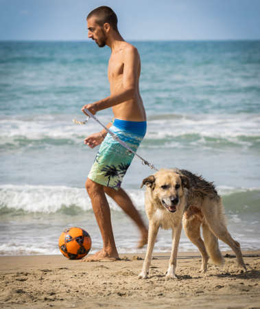 Bufalara, Italy - Septembre 02, 2020, Young man and large shepherd dog playing with bright ball on Bufalara beach in province of Latina in Italy during the second wave of coronavirus (covid 19)のeditorial素材
