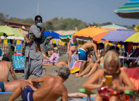 Marina di San Nikola, Italy - September 05, 2021, African merchant with beads and other jewelry wears a medical mask on the beach among crowds of people without masks in Marina di San Nicola, Italyのeditorial素材