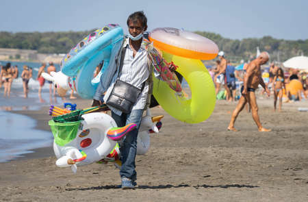 Marina di San Nikola, Italy - September 05, 2021, Bangladeshi inflatable toy dealer wears a medical mask on the beach among crowds of unmasked people in Marina di San Nicola, Italy.のeditorial素材