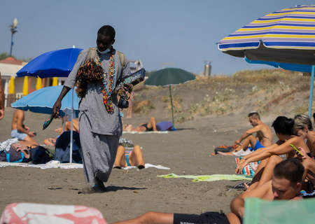 Marina di San Nikola, Italy - September 05, 2021, African merchant with beads and other jewelry wears a medical mask on the beach among crowds of people without masks in Marina di San Nicola, Italyのeditorial素材