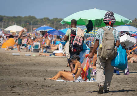 Marina di San Nikola, Italy - September 05, 2021, Hat vendor on the beach among crowds of unmasked people in Marina di San Nicola, Italy.のeditorial素材