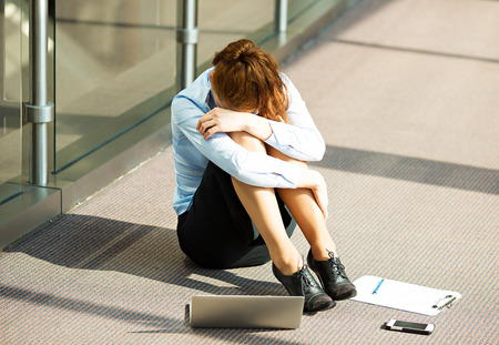 Closeup portrait young depressed, sad stressed, crying businesswoman sitting on floor in a hall of corporate building, company office, covering her face, with laptop cell phone, documents lying aroundの写真素材