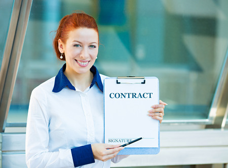 Closeup portrait smiling young business woman, company employee holding contract document pointing with pen at space for signature isolated corporate office background Positive face expression emotionの写真素材