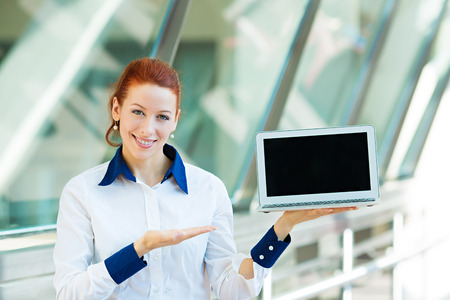 Closeup portrait smiling business woman holding laptop notebook computer, showing something on screen isolated background corporate office windows. Positive facial expression, emotion, attitudeの写真素材