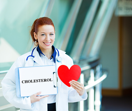 Closeup portrait happy smiling female health care professional, woman family doctor, cardiologist with stethoscope holding sign cholesterol, heart isolated hospital hallway background. Patient planの写真素材