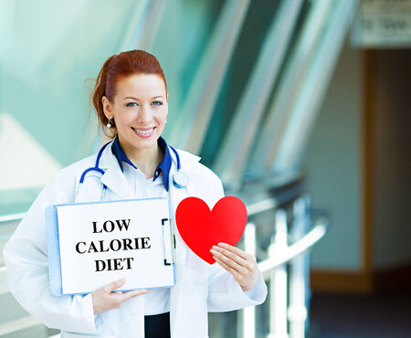 Closeup portrait happy smiling female health care professional woman family doctor, cardiologist with stethoscope holding sign low calorie diet heart isolated hospital hallway background. Patient planの写真素材