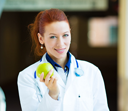 Closeup portrait smiling happy health care professional, doctor, nurse in lab coat offering green apple, isolated background hospital hallway. A day keeps doctor away concept. Nutrition, healthy dietの写真素材