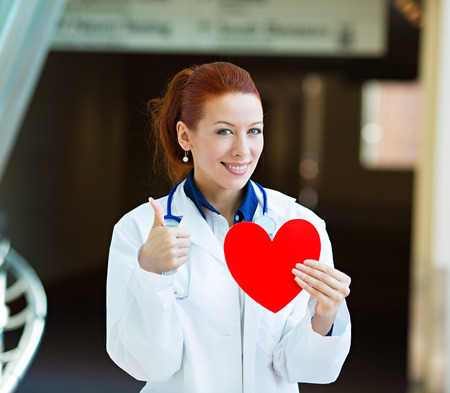 Closeup portrait happy smiling female health care professional, woman family doctor cardiologist with stethoscope holding red heart, giving thumbs up isolated hospital hallway background. Patient planの写真素材