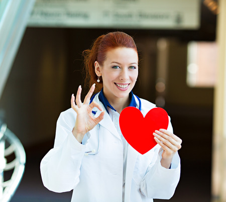 Closeup portrait happy smiling female health care professional, woman family doctor, cardiologist with stethoscope holding red heart, giving ok sign isolated hospital hallway background. Patient planの写真素材