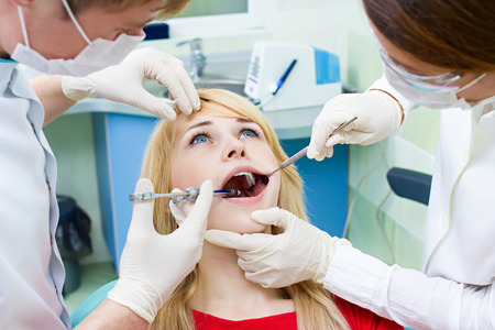 Closeup portrait young female, woman patient sitting in dentist chair, office with wide open mouth getting painful anesthetic injection done by doctor, his assistant isolated background clinic officeの写真素材
