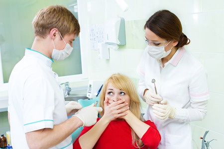 Closeup portrait young terrified girl, woman scared at dentist visit, siting in chair, covering her mouth, doesn't want dental procedure, drilling, tooth extraction, isolated clinic office backgroundの写真素材