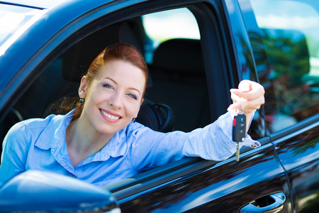 Closeup portrait happy, smiling, young, attractive woman, buyer sitting in her new black car showing keys isolated city street dealership lot background. Personal transportation, auto purchase conceptの写真素材