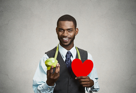 Closeup portrait, headshot business man, corporate executive holding green apple, red heart measuring tape on neck isolated black grey background. Healthy life style concept. Positive face expressionの写真素材
