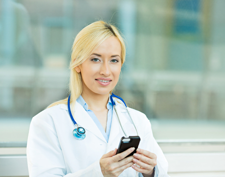 Closeup portrait young healthcare professional, doctor, nurse, dentist, researcher, physician assistant reading text sms, message on cellphone holding smart phone isolated hospital hallway  backgroundの写真素材