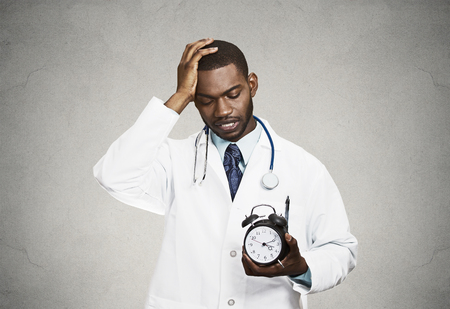 Closeup portrait overwhelmed with busy schedule health care professional doctor, nurse, dentist with stethoscope holding alarm clock running out of time having headache isolated black grey backgroundの写真素材