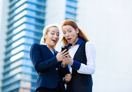 Closeup portrait happy, surprised business women, company partners receiving news on mobile phone, looking at smartphone reading text message celebrating success, isolated background corporate officeの写真素材