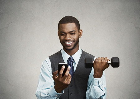 Closeup portrait happy, smiling business man reading good news on smart phone, holding mobile, lifting weight, dumbbell isolated black background. Human face expression, emotion, corporate executiveの写真素材