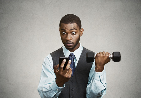 Closeup portrait shocked, surprised business man reading bad news on smart phone holding mobile, lifting weight, dumbbell isolated black background. Human face expression, emotion, corporate executiveの写真素材