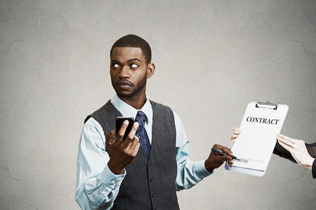 Closeup portrait annoyed skeptical sneaky businessman signing contract without looking at document, corporate executive holding mobile smart phone isolated grey black background. Human face expressionの写真素材