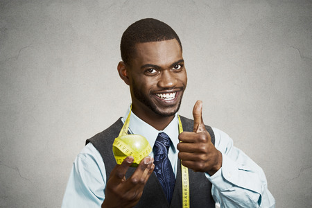 Closeup portrait, headshot business man, corporate executive holding green apple giving thumbs up measuring tape on neck isolated black background. Healthy life style concept. Positive face expressionの写真素材