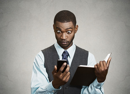 Closeup portrait surprised shocked business man executive reading breaking news on smart phone holding book isolated black grey background の写真素材
