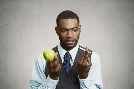 Closeup portrait headshot corporate executive businessman trying decide on healthy life choices, holding craving cigarette versus green apple isolated black background. Face expression body languageの写真素材