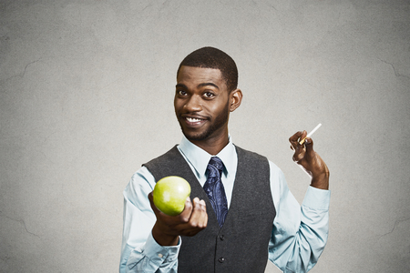 Closeup portrait, headshot handsome, happy, smiling, young business man says no to cigarette, offers, makes healthy life choice, holding green apple isolated black background. Human face expressionsの写真素材