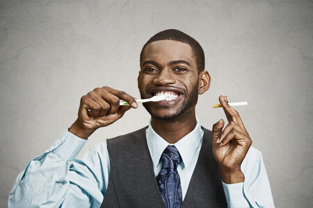 Closeup portrait, headshot  young business man, company, corporate executive holding toothbrush, cigarette, isolated black background. Smoker breath concept. Face expression, perception. Oral hygieneの写真素材