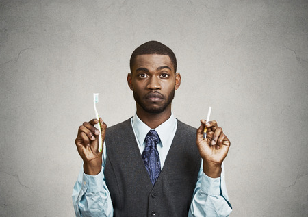 Closeup portrait, headshot  young business man, company, corporate executive holding toothbrush, cigarette, isolated black background. Smoker breath concept. Face expression, perception. Oral hygieneの写真素材