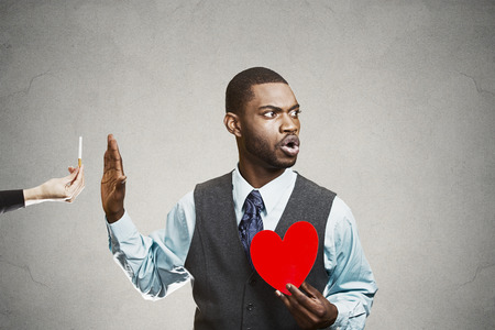 Closeup portrait, headshot handsome young business man says no to cigarette offered by person, stop hand gesture, holding heart isolated black  background. Healthy life choices. Human face expressionの写真素材