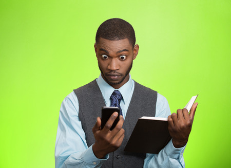 Closeup portrait surprised shocked business man executive reading breaking news on smart phone holding book isolated green background.  の写真素材