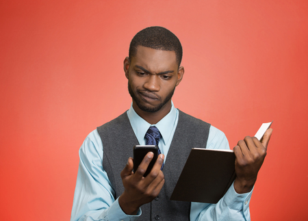 Closeup portrait confused, skeptical business man, executive reading news on smart phone, holding book isolated red background. Human face expression, emotion, body language, corporate lifestyle の写真素材