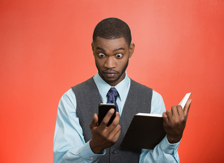 Closeup portrait surprised shocked business man executive reading breaking news on smart phone holding book isolated red background. Human face expression, emotion, corporate company lifestyle の写真素材