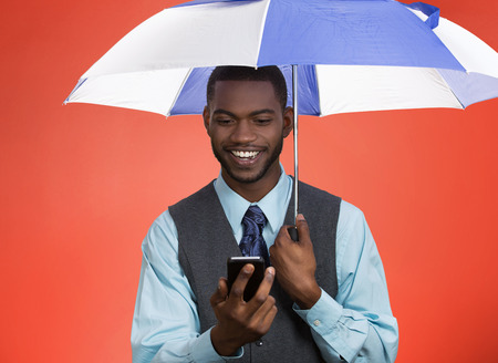 Closeup portrait happy, smiling corporate business man, executive reading message news on smart phone holding umbrella, protected from rain isolated red background. Human face expression, emotion の写真素材
