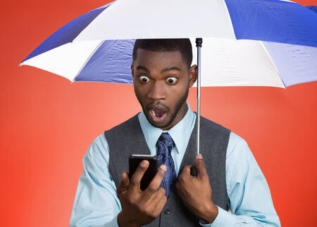Closeup portrait shocked, surprised business man, corporate executive reading bad, breaking news on smart phone holding umbrella protected from rain isolated red background. Face expression, emotion の写真素材