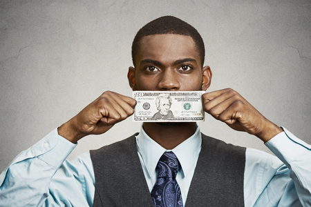 Closeup portrait handsome young corrupt corporate man in blue shirt holding twenty dollar bill to mouth, isolated grey background. Bribery concept in politics, business, diplomacy. Life perceptionの写真素材