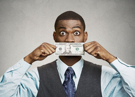 Closeup portrait shocked, surprised young corporate man in blue shirt holding twenty dollar bill to mouth, isolated grey background. Bribery concept in politics, business, diplomacy. Life perceptionの写真素材