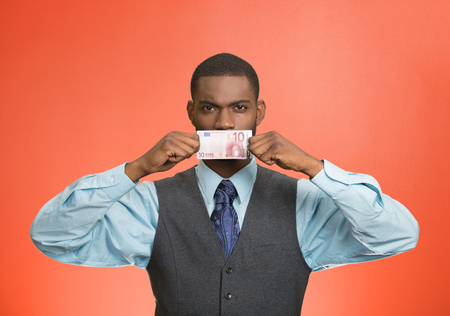 Closeup portrait handsome young corrupt corporate man in blue shirt holding euro bill currency to his mouth, isolated red background. Bribery concept in politics, business, diplomacy. Life perceptionの写真素材