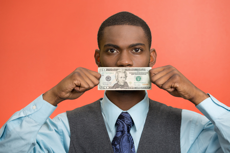 Closeup portrait handsome young corrupt corporate man in blue shirt holding twenty dollar bill to mouth, isolated red background. Bribery concept in politics, business, diplomacy. Life perceptionの写真素材