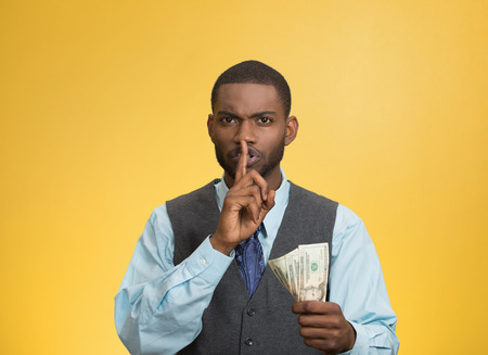 Closeup portrait handsome corrupt guy businessman holding dollar bill in hand showing shhh sign finger to lips isolated yellow background. Bribery concept politics, business diplomacy. Face expressionの写真素材